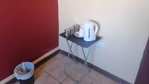 Una pequeña mesa con una tetera, tazas y recipientes, un cubo de basura y un enchufe en la pared.