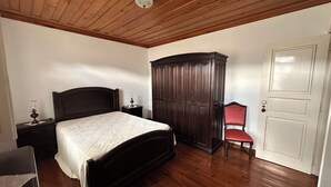 A bedroom with a wooden ceiling, a bed with a headboard, a wardrobe, a chair, and a door. - Casa João Fidalgo (Arcos de Valdevez)