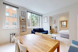 A modern living room with a wooden dining table, white chairs, a blue sofa, and a small white table. - City Centre 2BR Flat 7-Min Walk to British Museum (London)