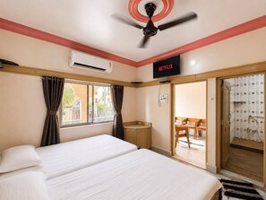 A hotel room with a bed, an air conditioning unit, a ceiling fan, a television displaying 'NETFLIX', and a view of the outside through a window. - Evergreen Bungalows (Dwarka)