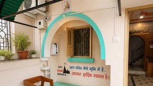Una habitación con una mesa y sillas de madera, una planta en maceta e inscripción religiosa en la pared.