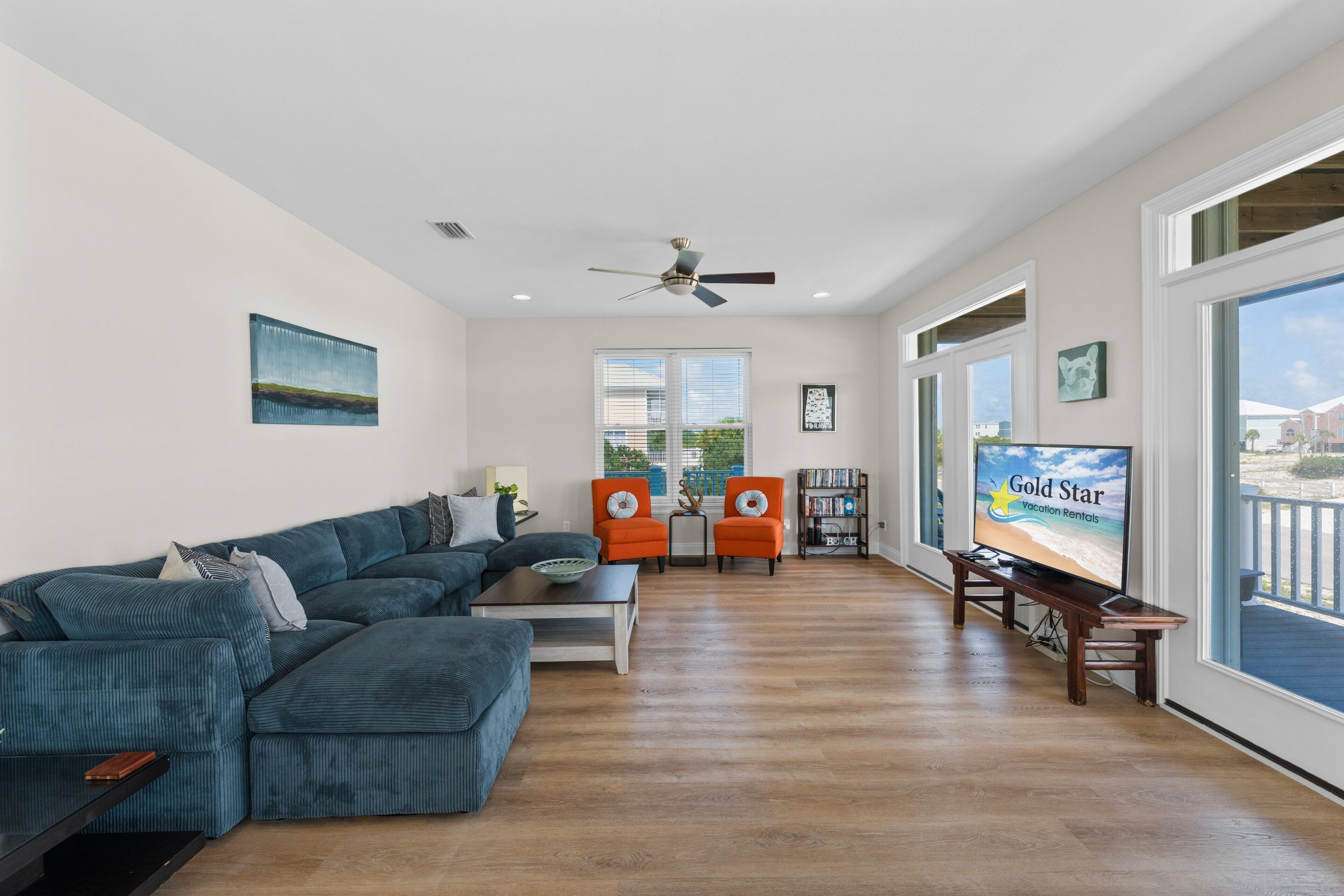 A spacious living room with a large sectional sofa, a coffee table, and a television on a stand. There are two armchairs and a bookshelf.