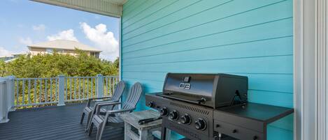 A covered outdoor area with a charcoal grill, two Adirondack chairs, and a view of a building in the background.