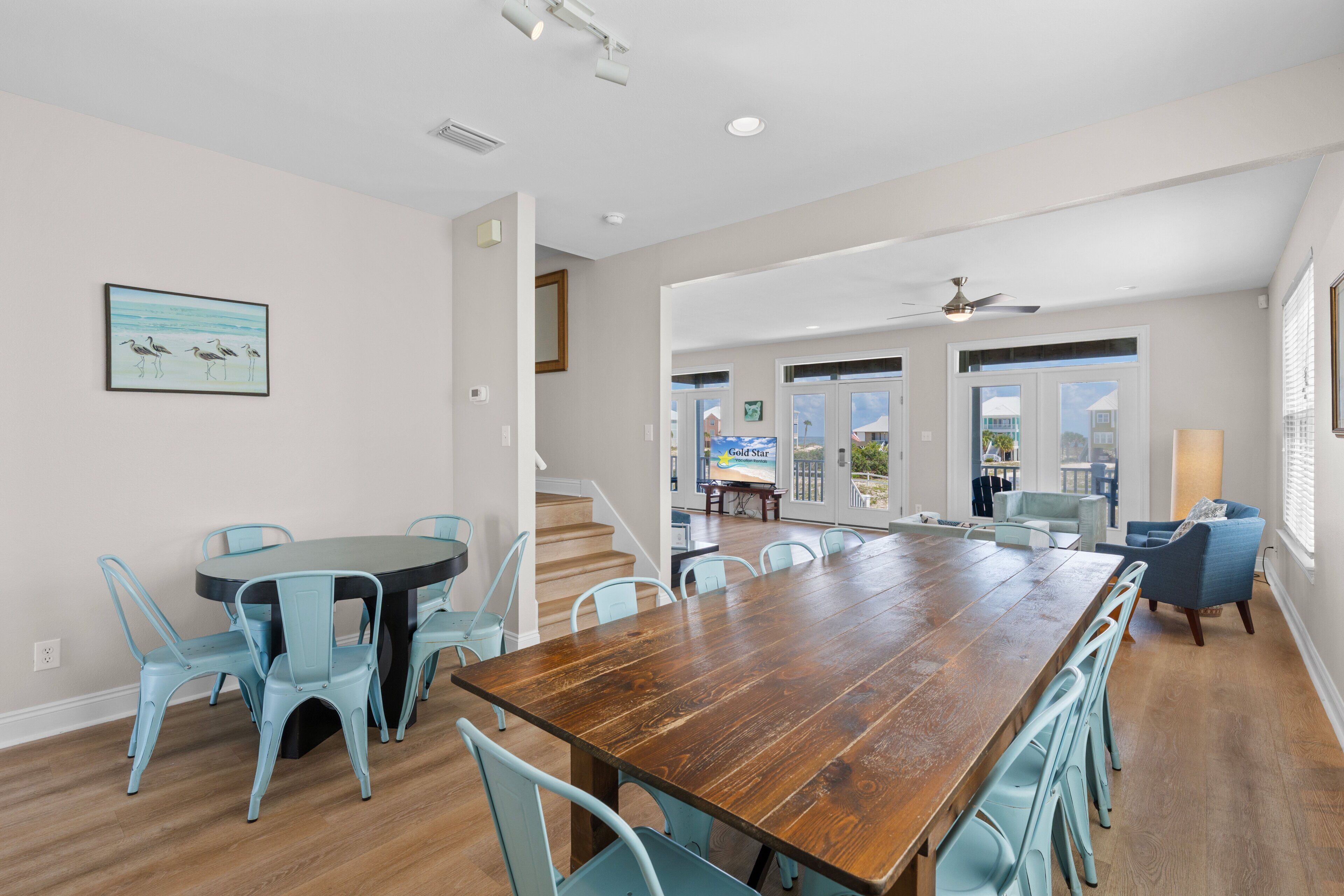 Un vaste espace à manger avec une grande table en bois et des chaises bleu clair, un escalier menant à un balcon et une vue sur l’océan.