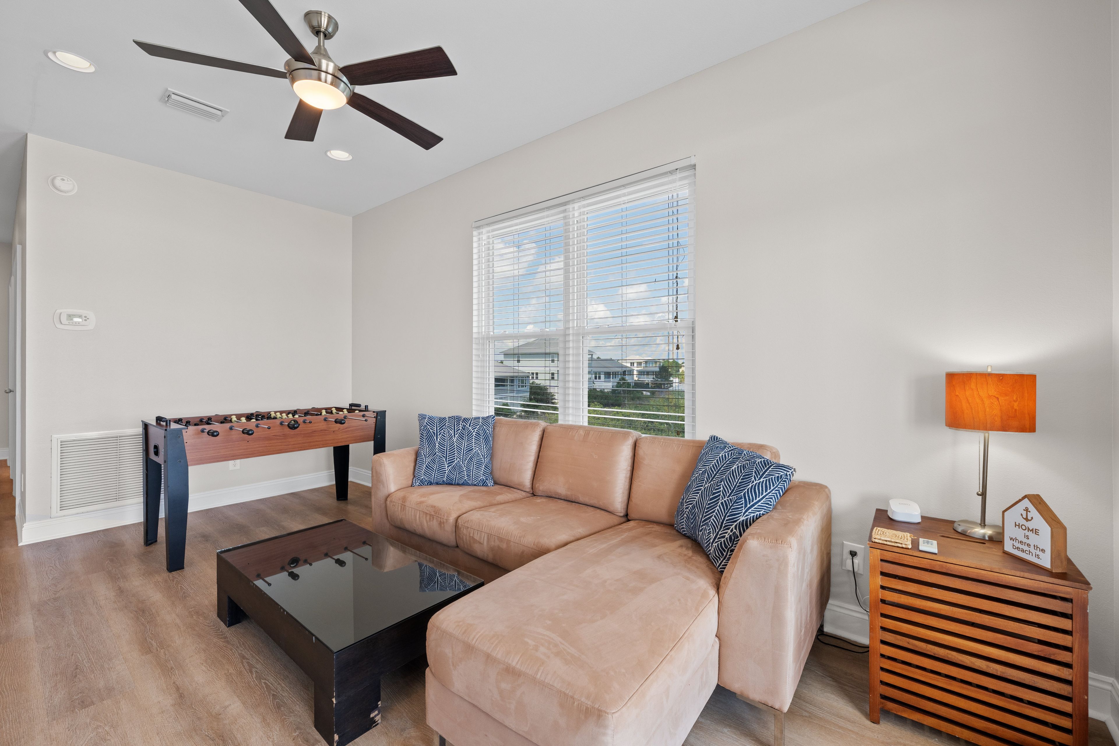 A living room with a sofa, a foosball table, a coffee table, a ceiling fan, and a window with blinds.