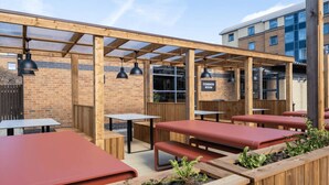 An outdoor seating area with wooden tables and benches, a common room sign, and a brick wall. - Student Only Clarence Dock (Leeds)