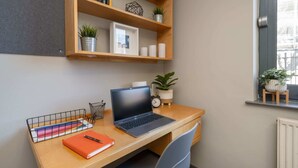 A workspace with a laptop, a notebook, and a pen on a wooden desk. There is a shelf above with decorative items and plants. - Student Only Clarence Dock (Leeds)