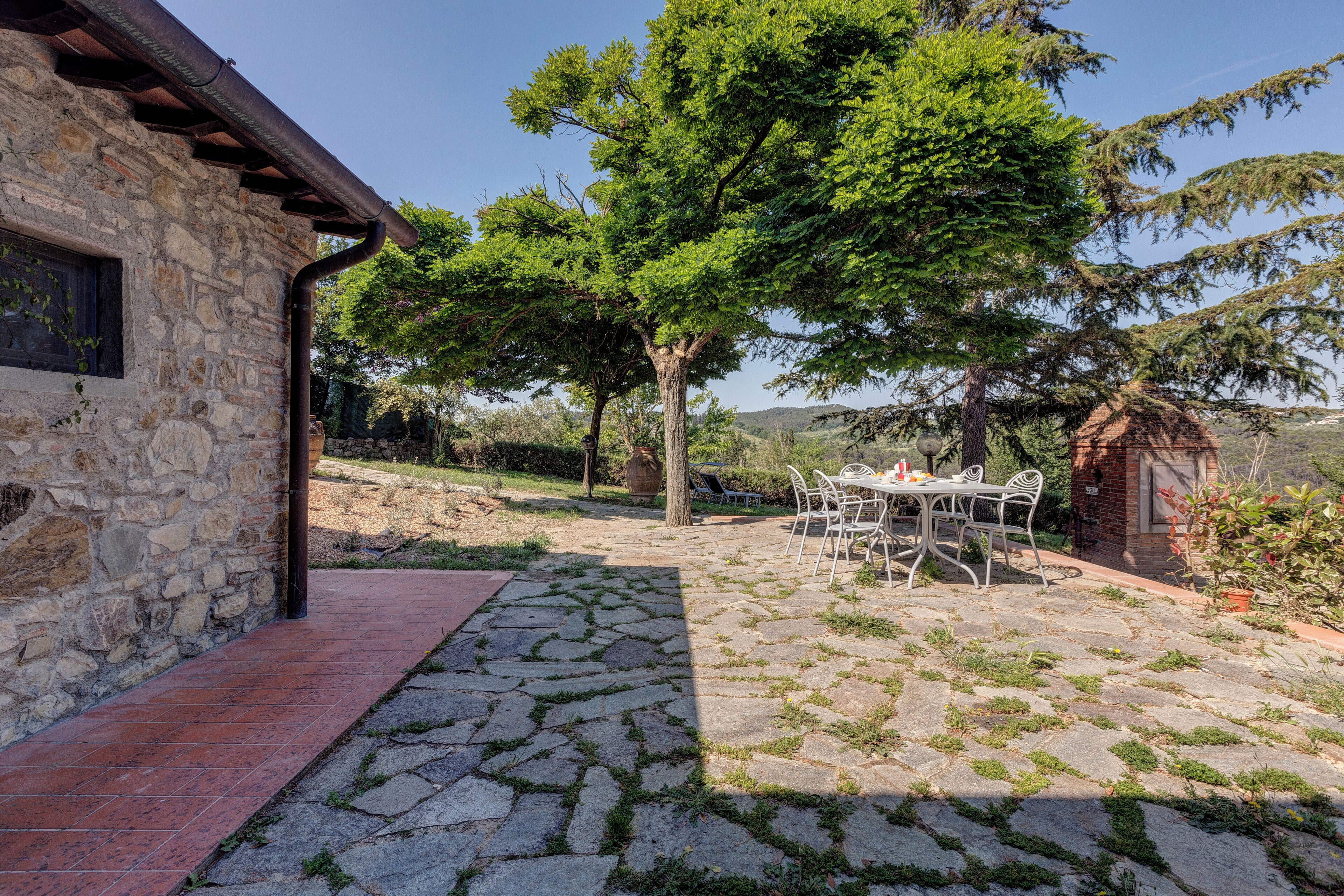 Un bâtiment en pierre avec une terrasse carrelée, une table entourée de chaises et un grand arbre en arrière-plan.