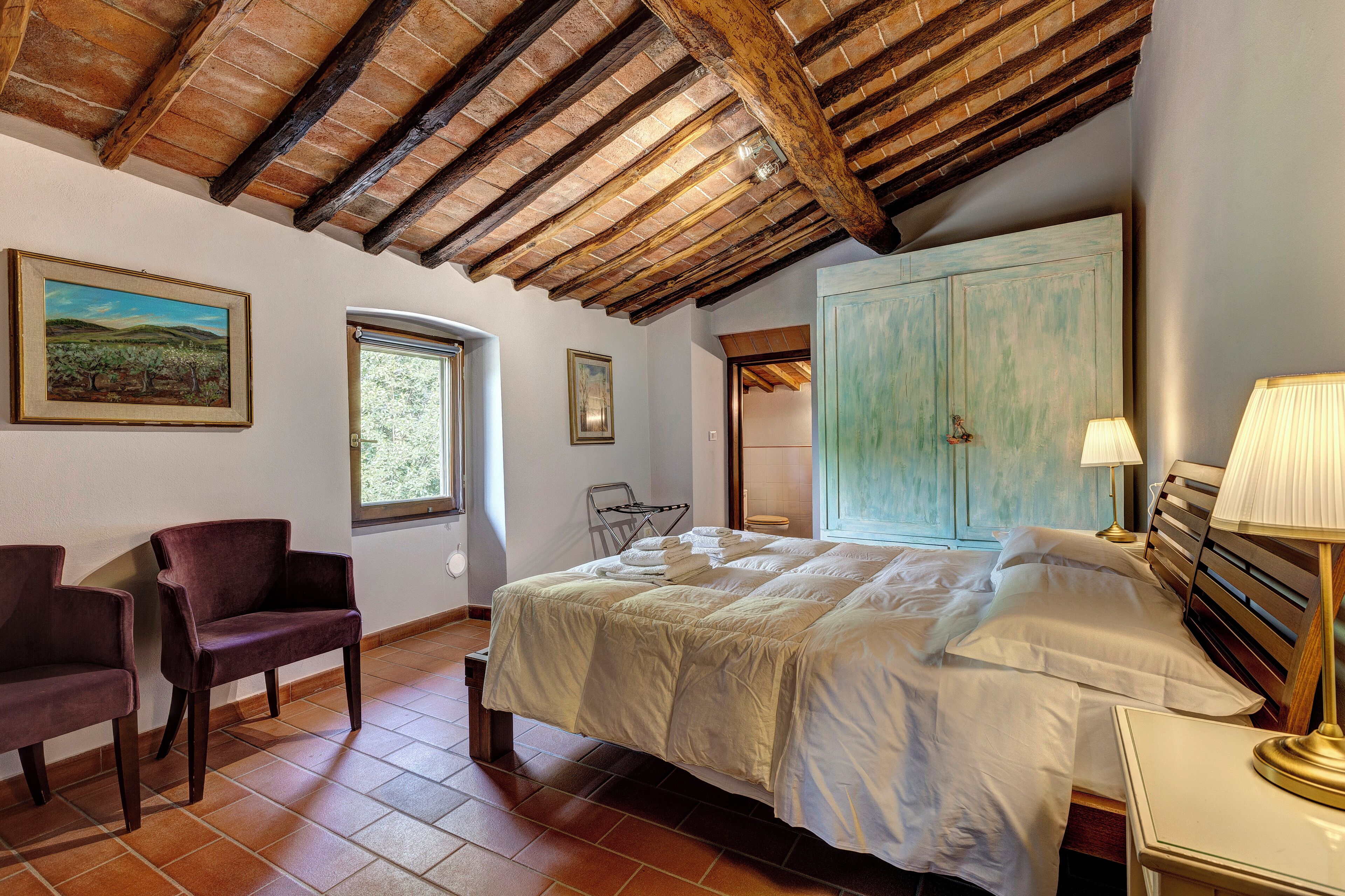 A bedroom with a large bed, a chair, a window, and a door leading to another room.