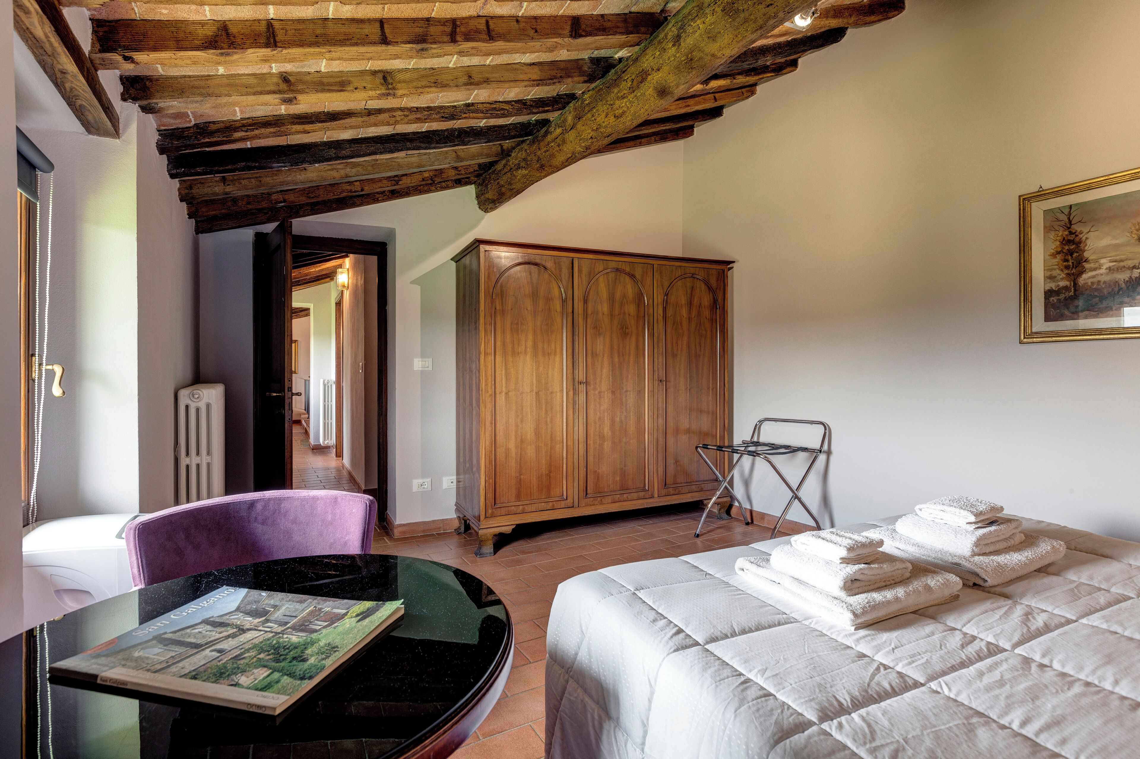 A bedroom with a bed, a wooden wardrobe, a chair, a table with a book, and a painting on the wall.