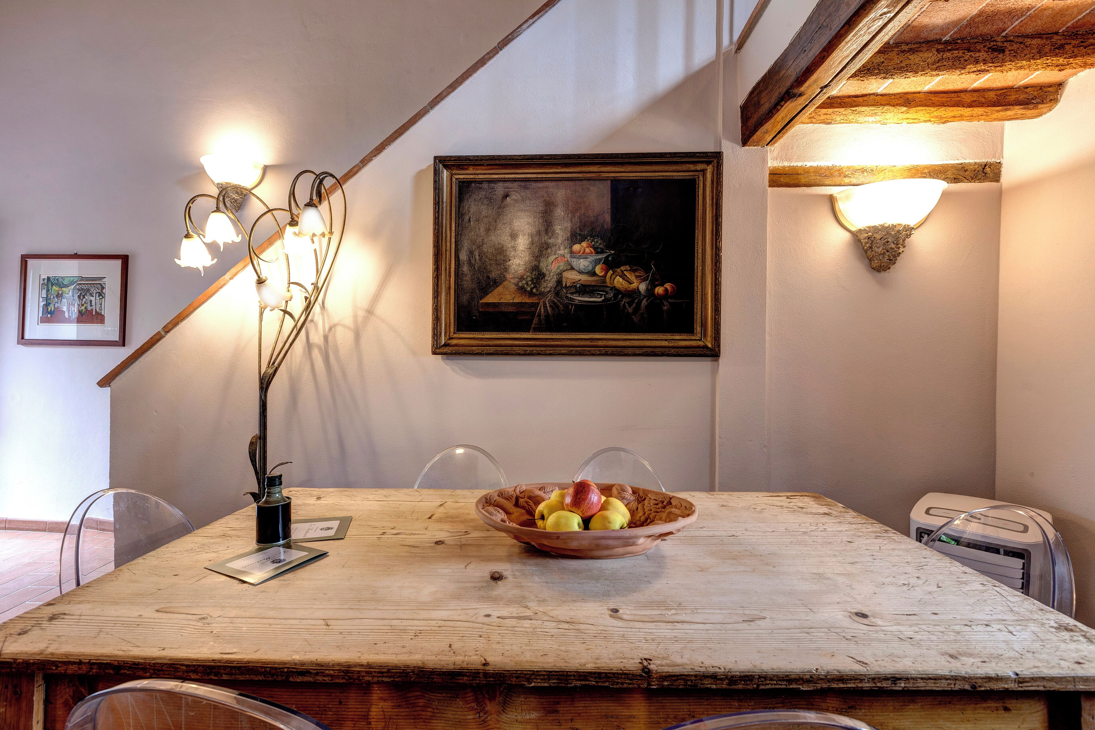 A dining area with a wooden table, a bowl of fruit, a framed painting, and wall-mounted lighting fixtures.