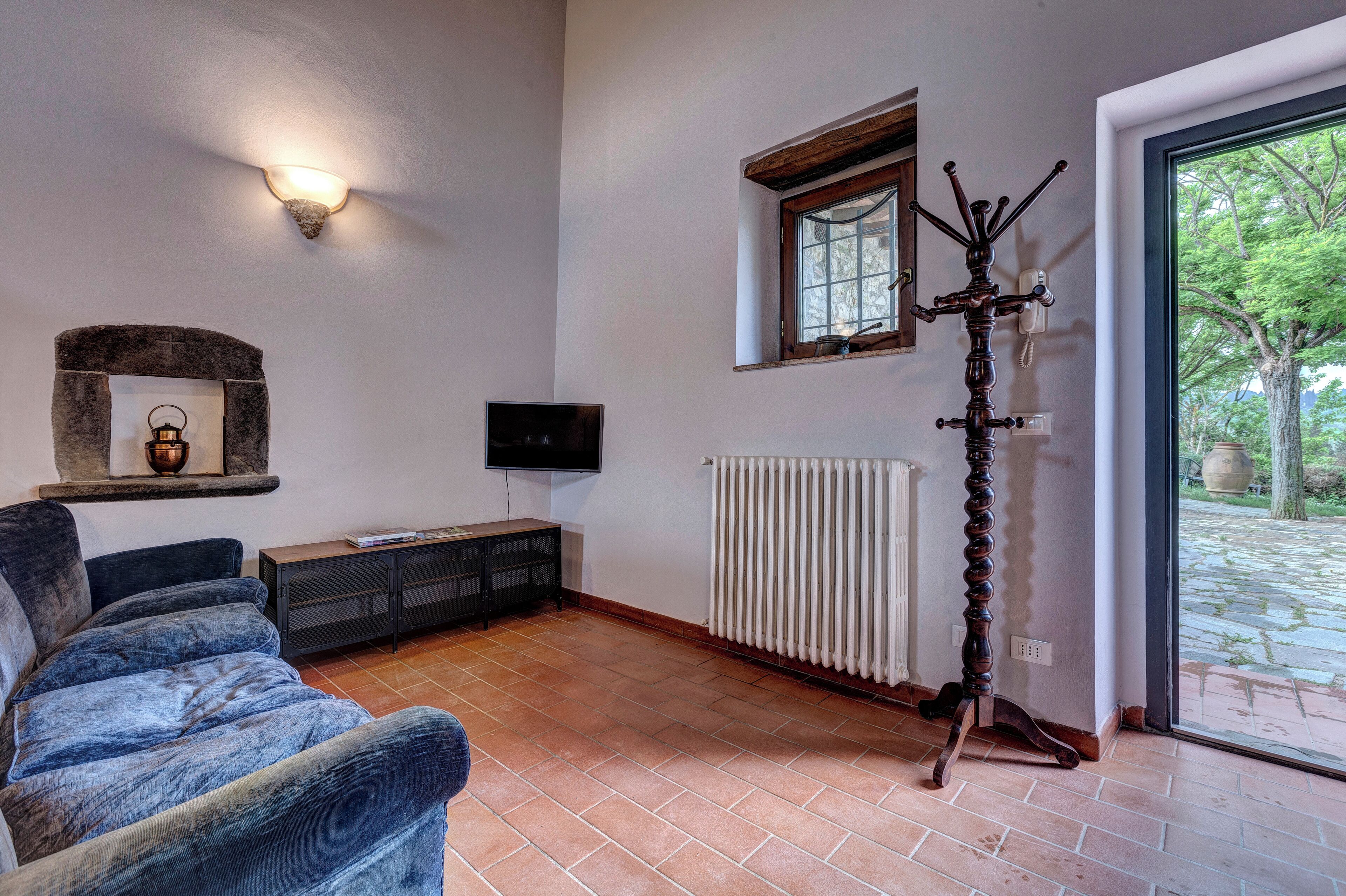 Een woonkamer met een bank, een televisie, een radiator en een raam met uitzicht op groen.