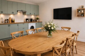A dining area with a round wooden table and six chairs, a washing machine, and a flat-screen TV mounted on the wall. - La Grande Maison Paris Sud (Vitry-sur-Seine)