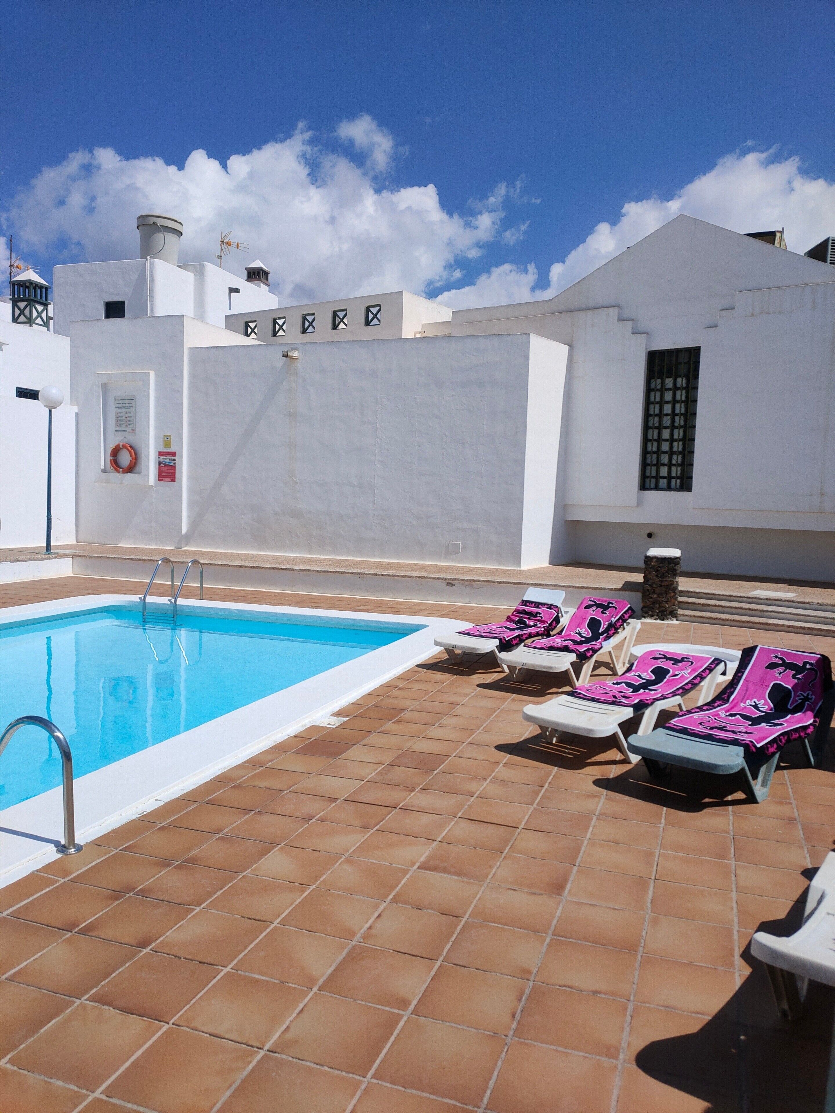 Una piscina con una scala, circondata da pareti bianche e un pavimento piastrellato. Sono presenti sedie a sdraio con asciugamani rosa.