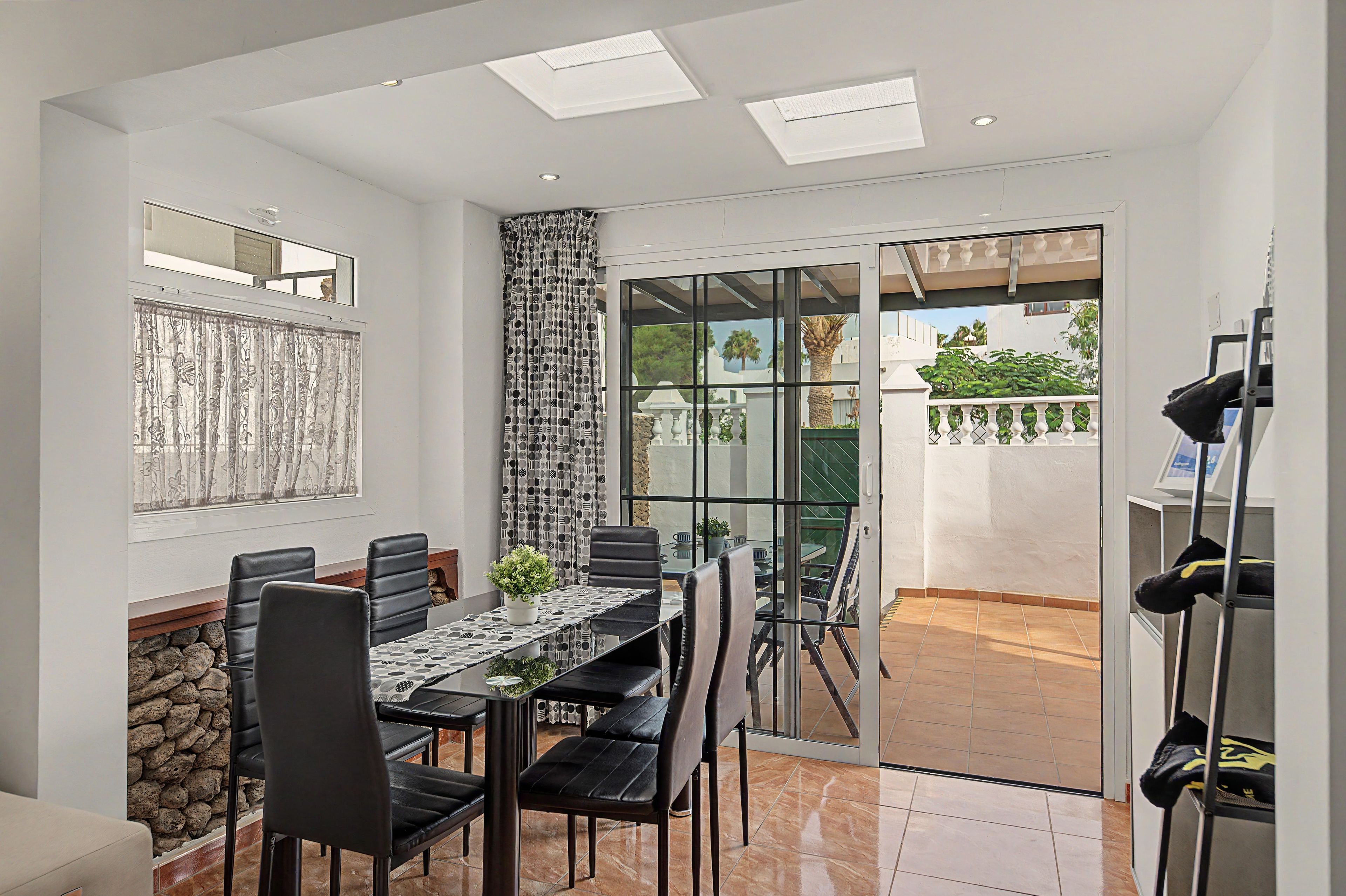 Uma área de refeições com mesa e cadeiras, um vaso de planta e vista através de uma porta de vidro para um espaço ao ar livre com piso ladrilhado e guarda-corpo.