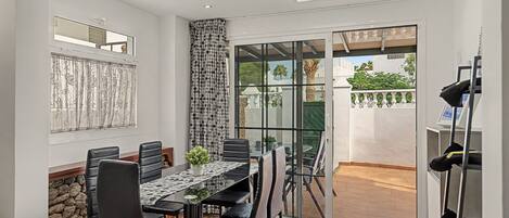 Uma área de refeições com mesa e cadeiras, um vaso de planta e vista através de uma porta de vidro para um espaço ao ar livre com piso ladrilhado e guarda-corpo.