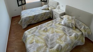 A room with two beds, a window with open shutters, a bedside table with a lamp, and a wall-mounted light. - Casa Princesa, cerca de la playa grande de Puerto del Carmen  (Tías)