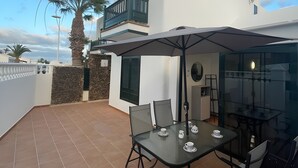 A modern outdoor dining area with a glass table and chairs, a white building with a balcony, and a palm tree. - Casa Princesa, cerca de la playa grande de Puerto del Carmen  (Tías)