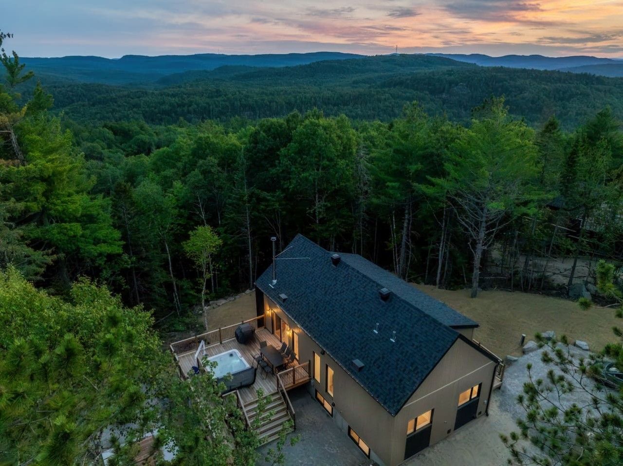 A modern two-story house with a deck and hot tub, surrounded by dense forest.