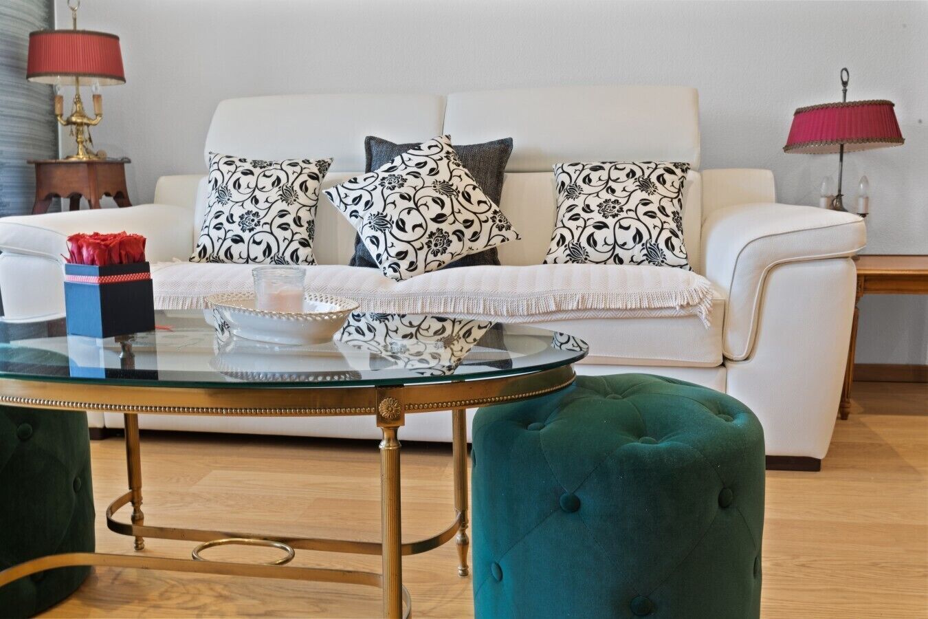 A living room with a white sofa, patterned cushions, a glass coffee table, and a teal ottoman.