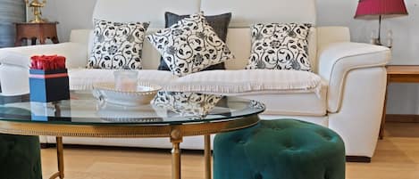 A living room with a white sofa, patterned cushions, a glass coffee table, and a teal ottoman.