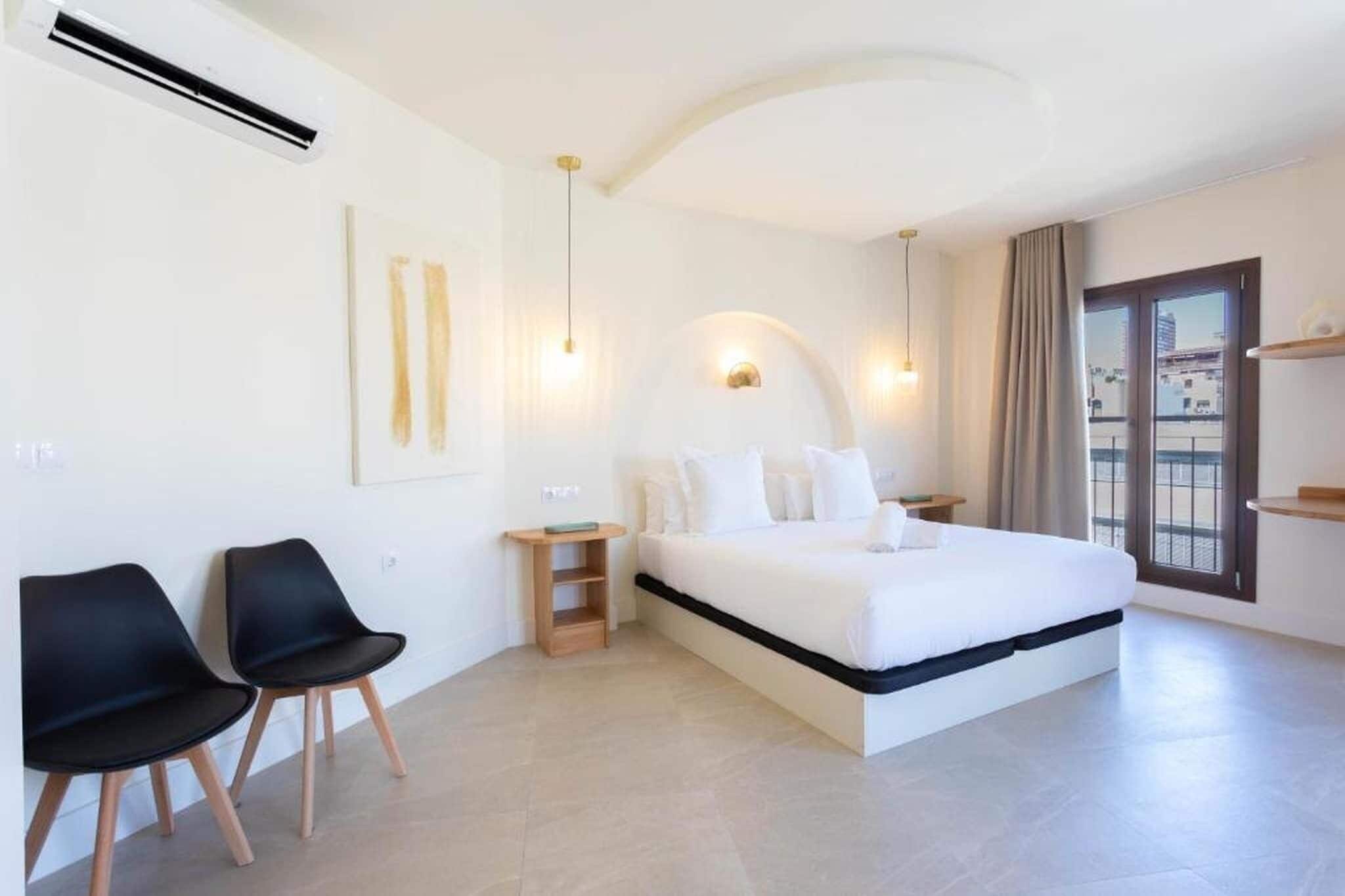Une chambre d’hôtel moderne avec un grand lit, deux chaises noires, une petite table d’appoint en bois et une grande fenêtre donnant sur un beau paysage.