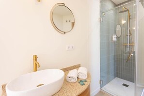 Ein Badezimmer mit einem großen weißen Waschbecken, goldenen Wasserhähnen, einem runden Spiegel und einer gläsernen Dusche.