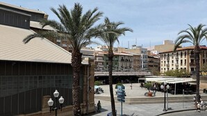 Una plaça pública amb palmeres, un mercat cobert i edificis amb balcons.
