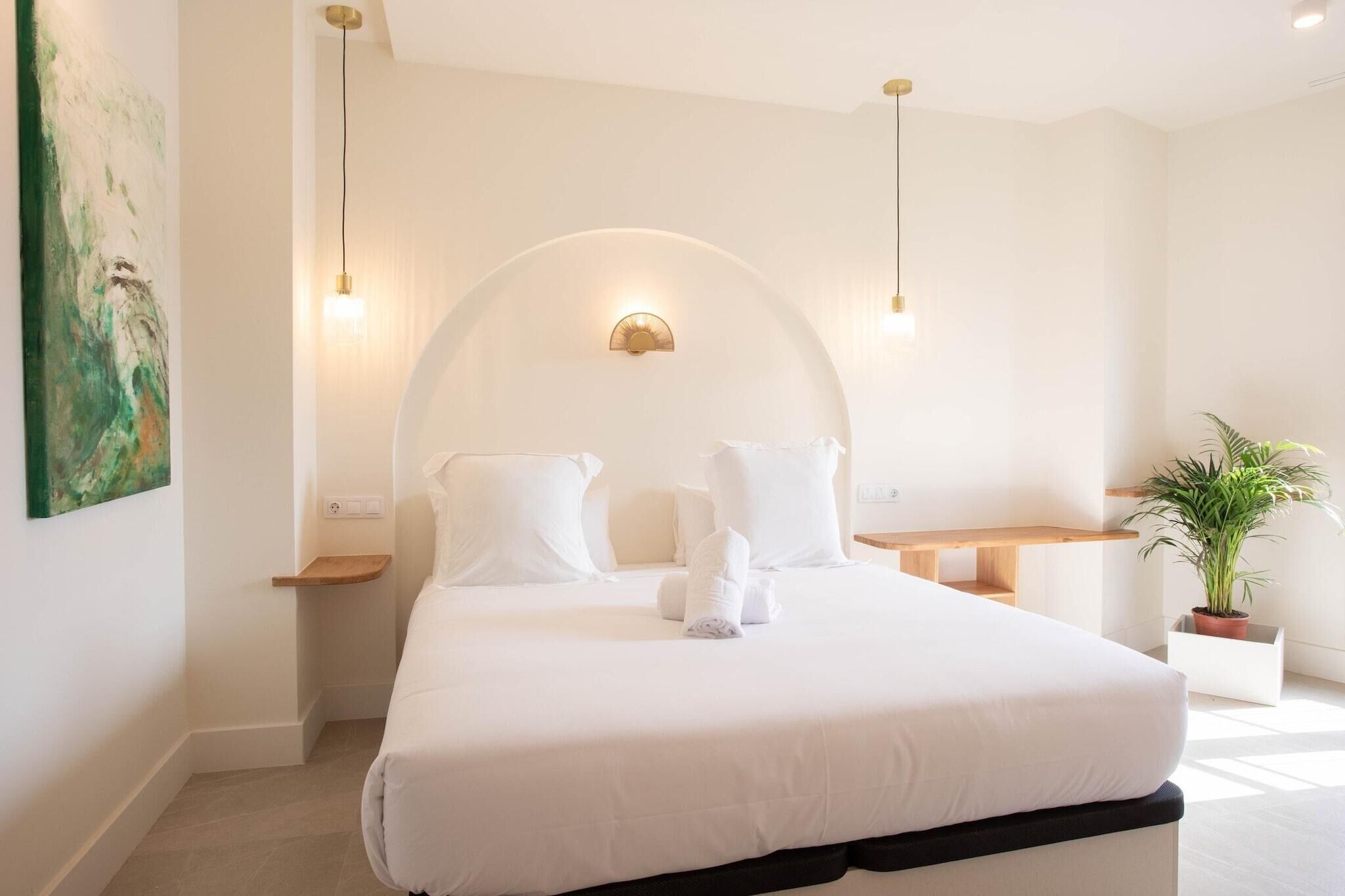A modern bedroom with a large bed, bedside tables, a wall-mounted light, and a potted plant.