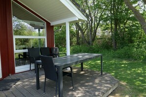 Ein überdachter Außenbereich zum Sitzen mit Tisch und Stühlen, umgeben von einer Holzterrasse und viel Grün.