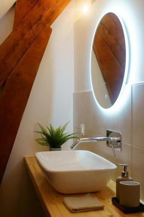 Une salle de bain avec un lavabo blanc, un miroir, une étagère en bois et une petite plante.