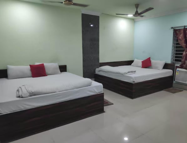 Habitación de hotel con dos camas, cada una con sábanas blancas y almohadas rojas, un ventilador de techo y una ventana con cortinas.