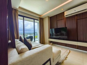 A modern living room with a large flat-screen TV, a sofa, and a view of the city through large windows. - Luxurious 2BR Condo With Ocean Views in Choeng Thale (Choeng Thale)