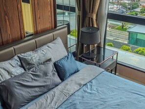 A modern hotel room with a large bed, a nightstand, a lamp, and a view of the city through large windows. - Luxurious 2BR Condo With Ocean Views in Choeng Thale (Choeng Thale)