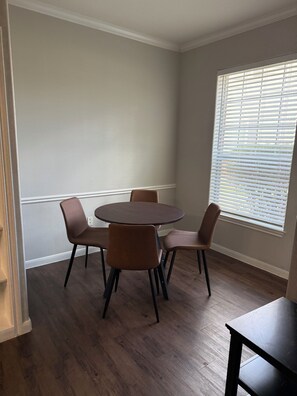 Ein Zimmer mit einem runden Tisch, vier Stühlen, einem Fenster mit Jalousien und Holzboden.