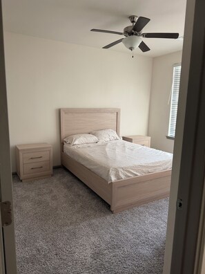 Quarto com cama, mesa de cabeceira, gaveteiro, ventoinha no teto e janela com persianas.