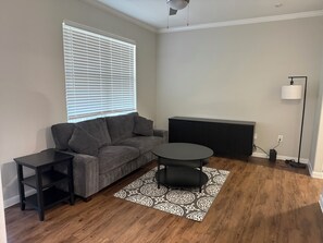 Ein Wohnzimmer mit einem grauen Sofa, einem runden Couchtisch, einem schwarzen Beistelltisch, einem gemusterten Teppich, einem schwarzen Heizkörper und einer Stehlampe.