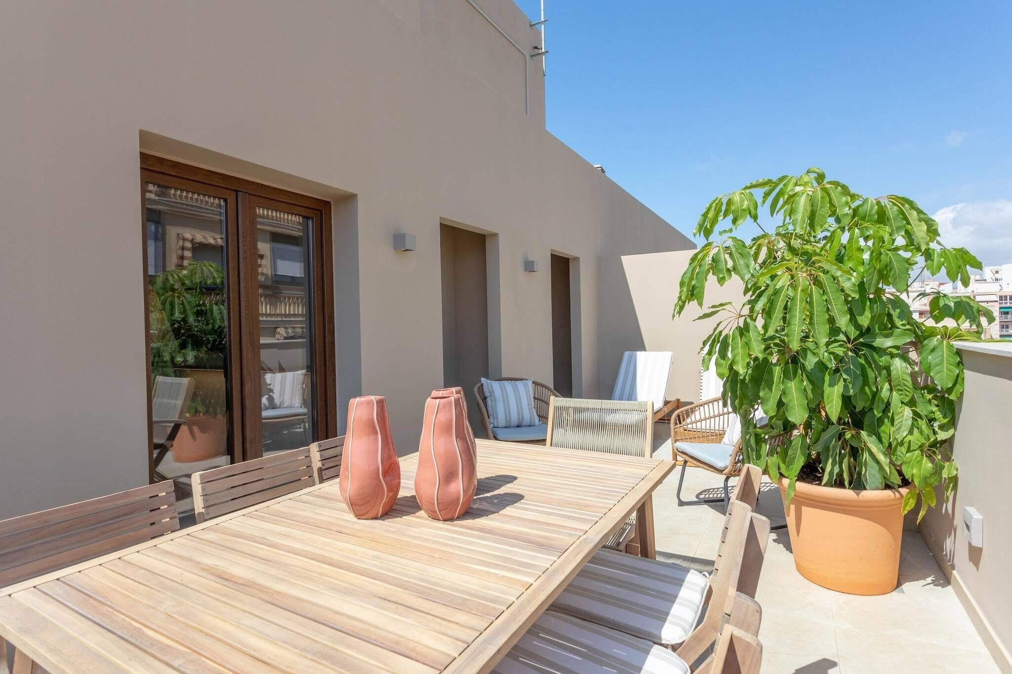 Un patio exterior con una mesa de madera, dos jarrones, sillas y una planta en maceta.