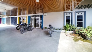 Um edifício com entrada coberta, duas motocicletas estacionadas em frente e um jardim com plantas e uma mangueira.