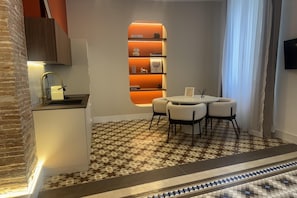 Habitación de hotel con área de cocina, una mesa de comedor con sillas, piso estampado y una estantería con libros.