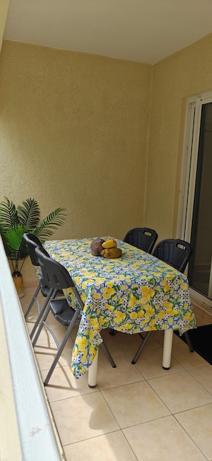 A dining table with a floral tablecloth, surrounded by chairs, in a room with a tiled floor and a sliding glass door. - Le Coin Grenadier - 2BR Apartment (Flic en Flac)
