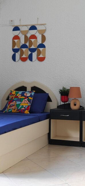 Un dormitorio con cama, almohada colorida, mesita de noche con lámpara, planta en maceta y un cuadro abstracto en la pared.