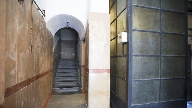 Um corredor estreito com escada que leva a uma porta com grade de metal.