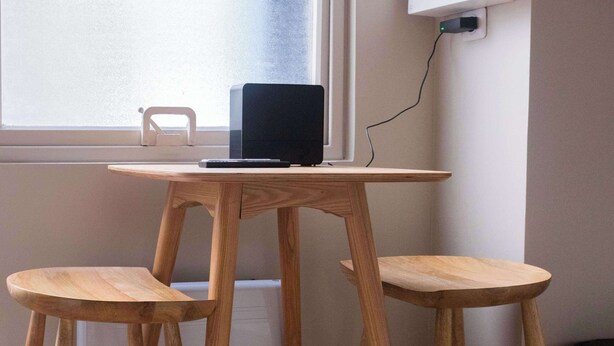Ein kleiner Holztisch mit einem Laptop und einem Stuhl, platziert in der Nähe eines Fensters mit einer Steckdose an der Wand.
