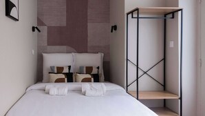 Ein ordentlich bezogenes Bett mit weißer Bettwäsche und vielen Kissen, ein Nachttisch mit Handtuch sowie ein an der Wand befestigtes Regalsystem.