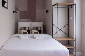 A neatly made bed with white bedding and multiple pillows, a bedside table with a towel, and a wall-mounted shelf unit. - Studio parisien cosy, proche Opéra/Saint-Lazare (Paris)