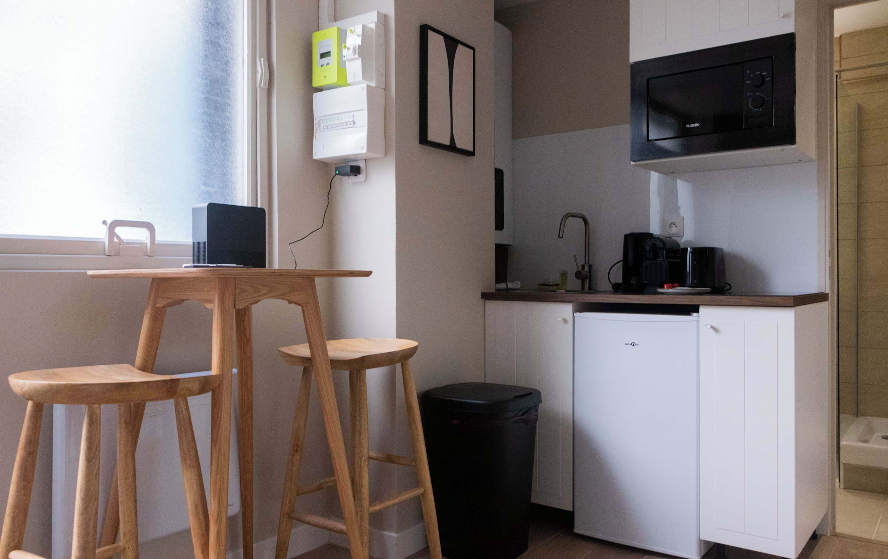 Een compacte keuken met een magnetron, koffiezetapparaat en een kleine koelkast. Er zijn twee houten krukken en een prullenbak.