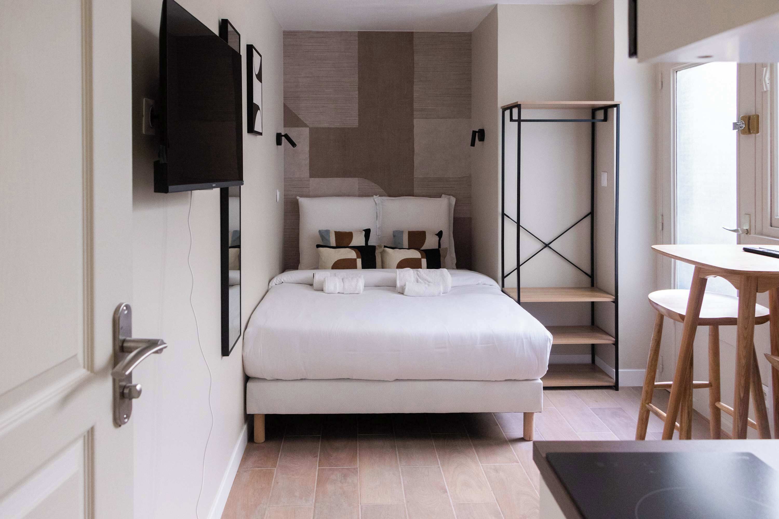 A modern bedroom with a bed, a TV mounted on the wall, a desk with a chair, and a small window.