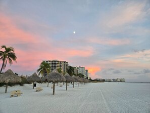 Una playa con sombrillas de paja y una hilera de edificios al fondo.