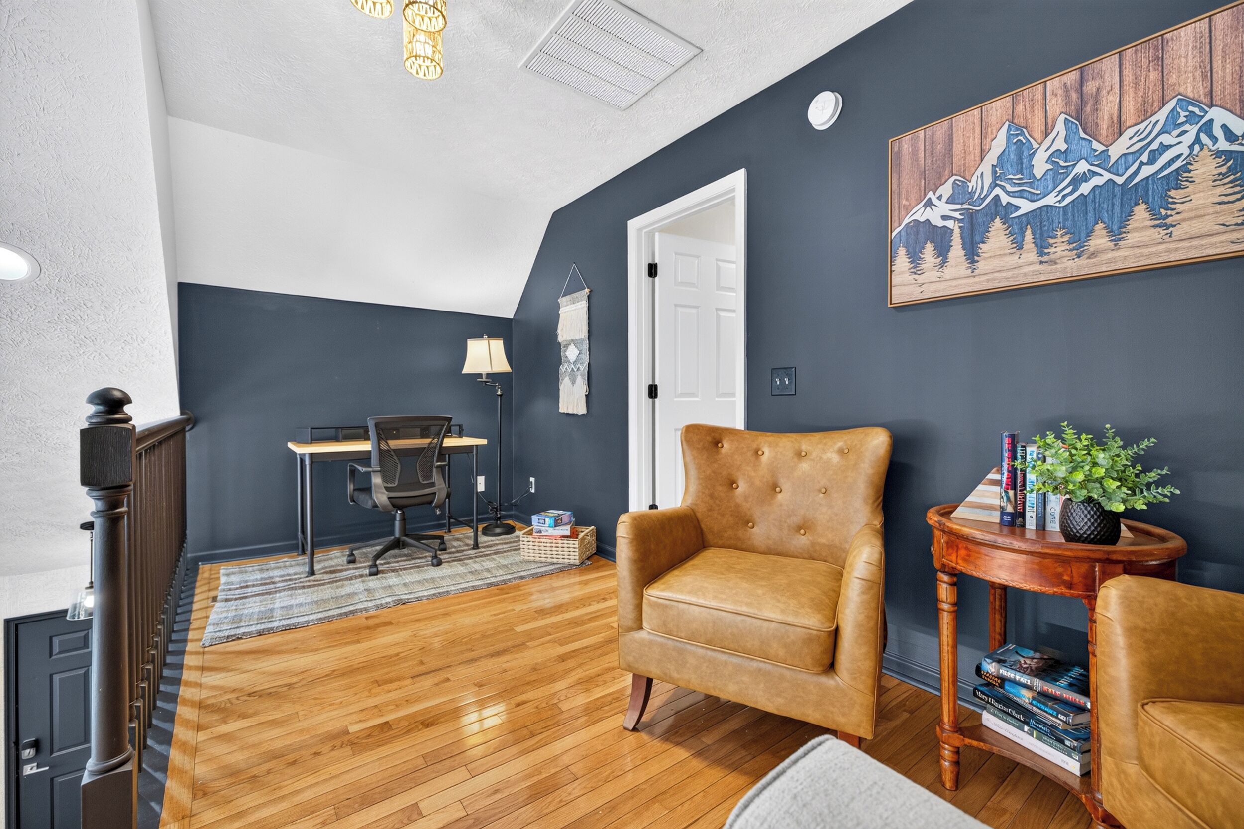 A room with a dark blue wall, a wooden floor, a leather armchair, a desk with a chair, a lamp, a small table with a plant, and a bookshelf.